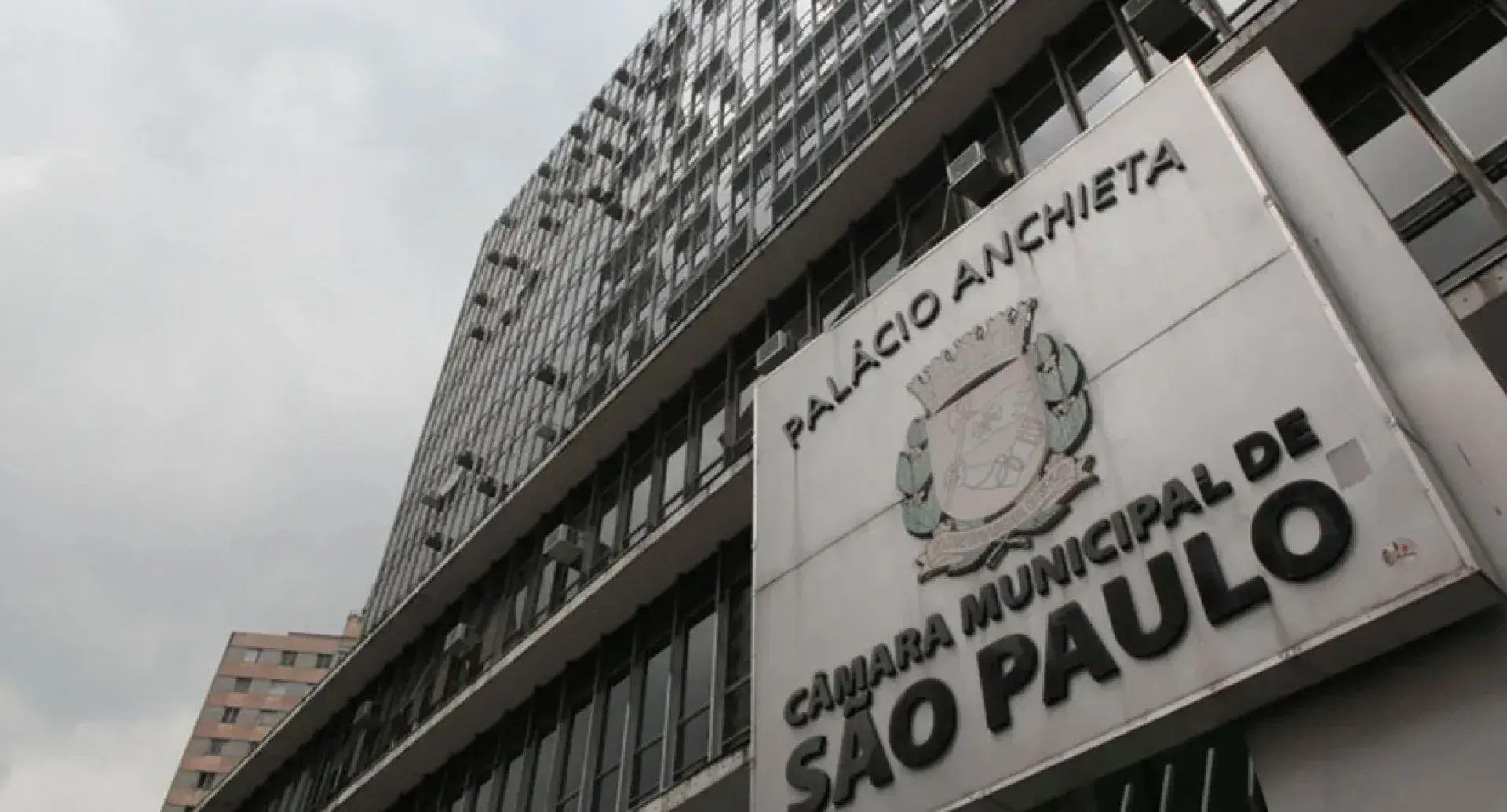 Fachada do Palácio Anchieta, sede da Câmara Municipal de São Paulo, vista de baixo, com a placa de identificação e o brasão da cidade em primeiro plano.
