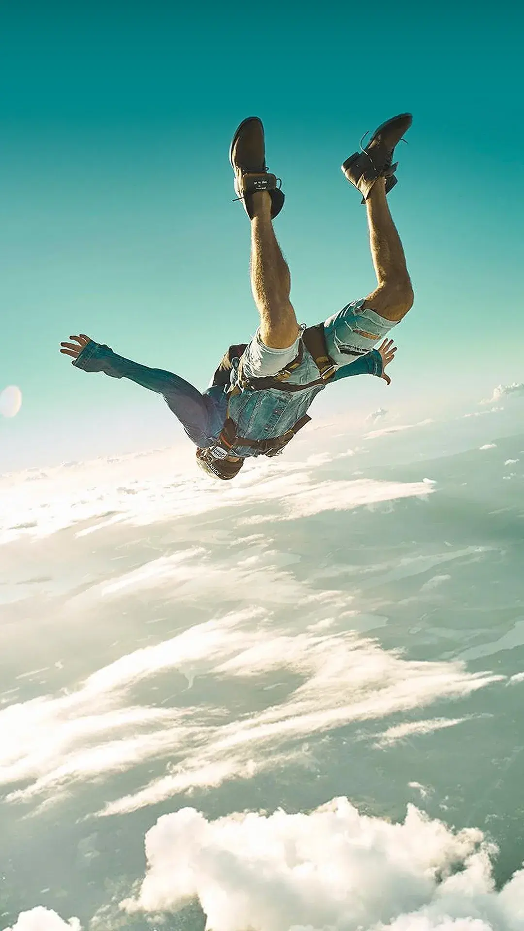 Paraquedista de cabeça para baixo em queda livre, de jaqueta e calça jeans da John John, com o mar de nuvens abaixo e o céu azul-turquesa ao fundo.