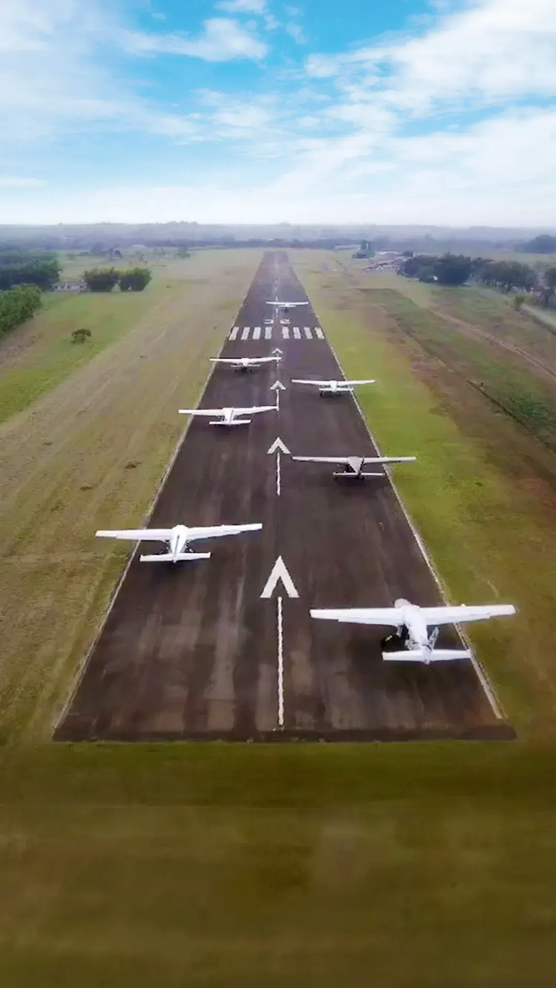 Seis aviões Cessna Caravan alinhados numa pista de aeroporto regional cercada por grama, vistos do alto.