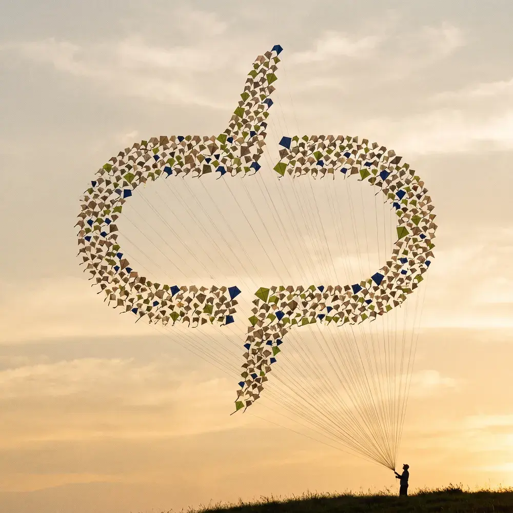 Céu de fim de tarde tomado por milhares de pipas, cada uma com um recorte e um padrão diferente. Vistas de baixo, todas juntas formam o símbolo da digigroup, e as linhas descem até uma única pessoa em silhueta na crista de um morro.